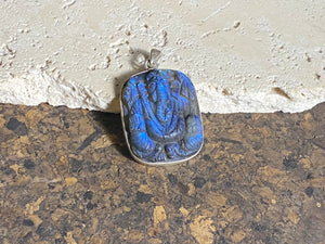 Hand carved square labradorite pendant depicting Ganesh, elephant headed god of wisdom, prosperity and new beginnings. This is a unique and individual piece, hand carved by an artisan in Jaipur, with a light blue luminosity that changes depending on the angle of lighting. Sterling silver frame and bail. The bail is generous enough to allow a large chain or cord to pass through.  This unisex labradorite pendant will suit both men and women.  Measurements: height 4.7 cm including bail, width 3 cm