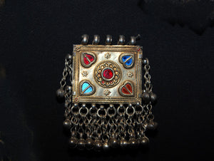 Antique Hunza Valley Uzbek pendant consisting of a flat, double skinned rectangular plaque with a central flower design surrounded by 4 stylised leaves, set with blue and red foil backed glass. Small bells hang in a fringe from the base of the pendant and the sides. Silver and faint gilding. Height 7.4 cm