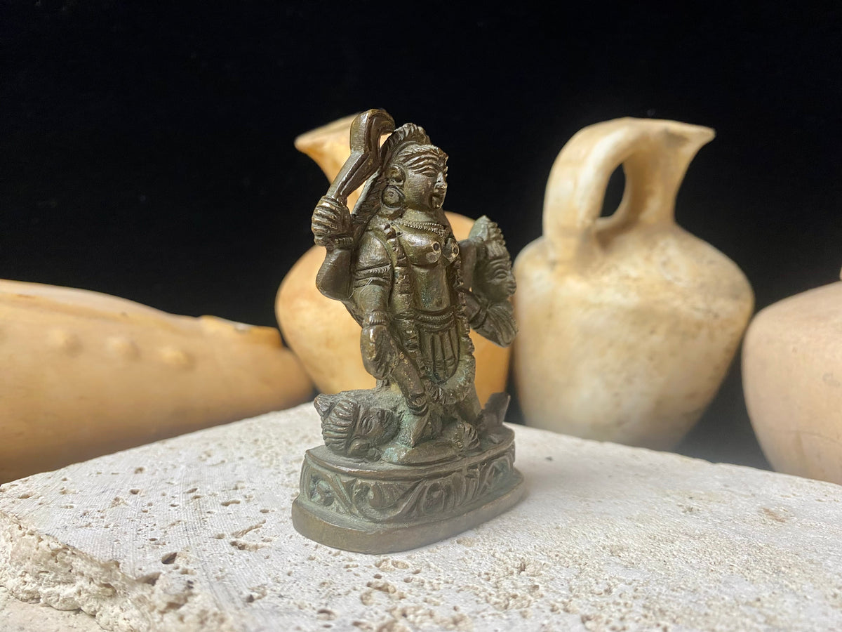 Kali with four arms, standing on Shiva, holding a curved sword, severed head and skull cup. Her fourth hand displays the gesture of blessing. She wears a garland of skulls around her neck and is covered by her long, wild, disheveled hair. This statue is approximately 80 years old (early 20th century). She has a fine and evenly dark patina.  Measurements: height 7.3 cm, width 5 cm.