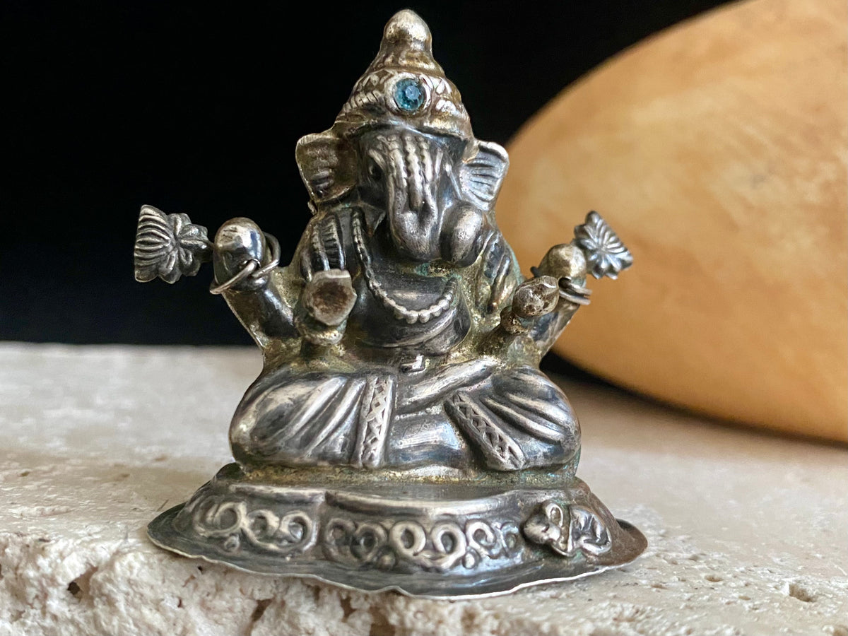 Small vintage Ganesh statue cast in silver, with right hand up in the gesture of protection and left hand holding his favourite ladoo sweet. His other hands hold lotus flowers. Lightweight but very finely cast with a small facet cut blue topaz in his forehead. 50 years old, southern India. 5 cm height