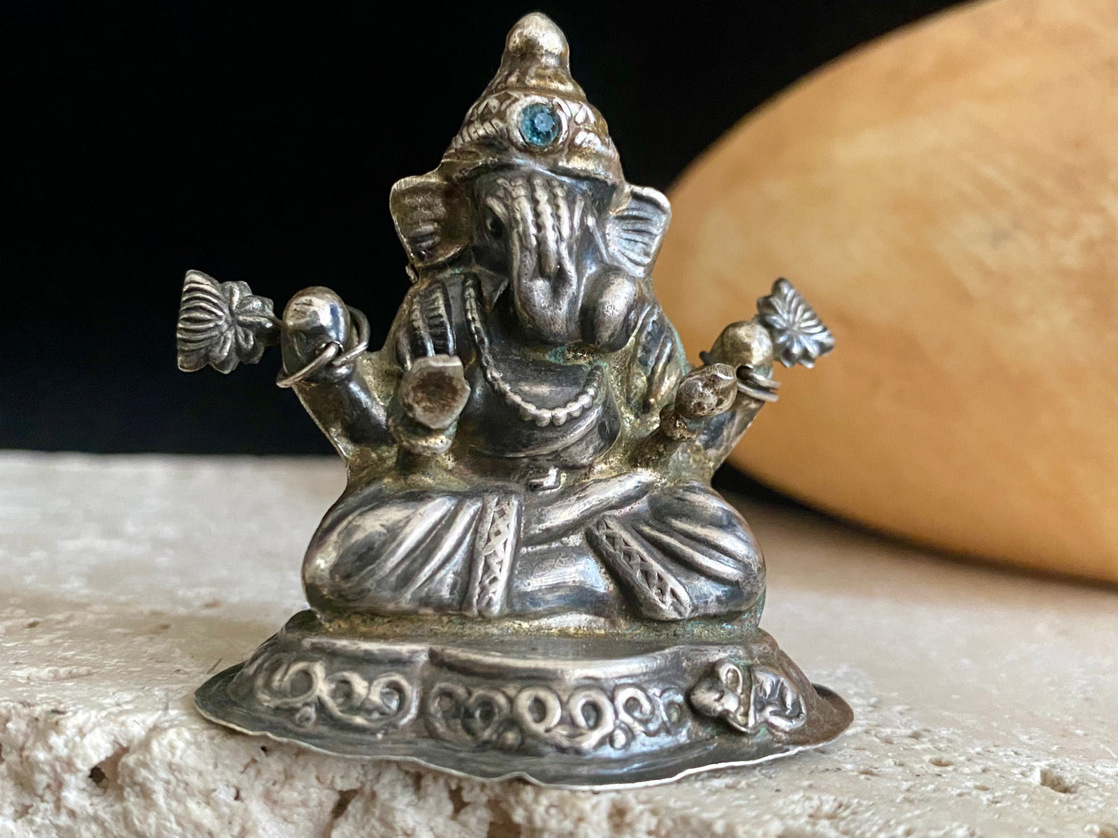 Small vintage Ganesh statue cast in silver, with right hand up in the gesture of protection and left hand holding his favourite ladoo sweet. His other hands hold lotus flowers. Lightweight but very finely cast with a small facet cut blue topaz in his forehead. 50 years old, southern India. 5 cm height