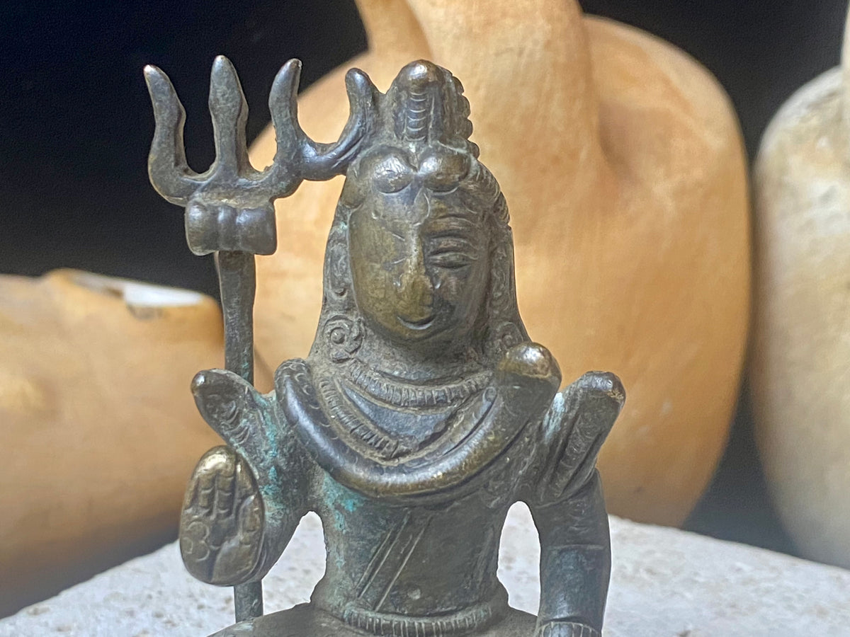 A small cast brass Shiva in serene seated style. This piece has some rubbing on the right hand side of the face and displays softened edges from decades of handling. It has a naturally darkened patina and is approximately 80 years old, or older. Gujarat, India. Measurements: Height 7.7 cm x width 5 cm x depth 3.5 cm