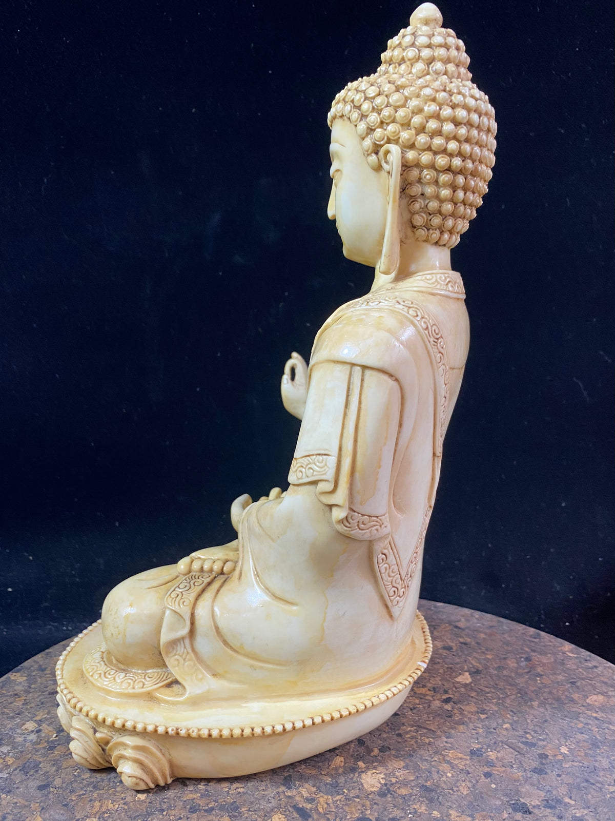 Amoghshiddhi Buddha cast in cream coloured resin. His right hand is held up facing outwards in the gesture of protection of fearlessness while his left hand sits in his lap in the gesture of meditation. Cast in solid resin, then hand finished to a very high standard. Measurements: height 26 cm, width 18 cm, depth 12 cm