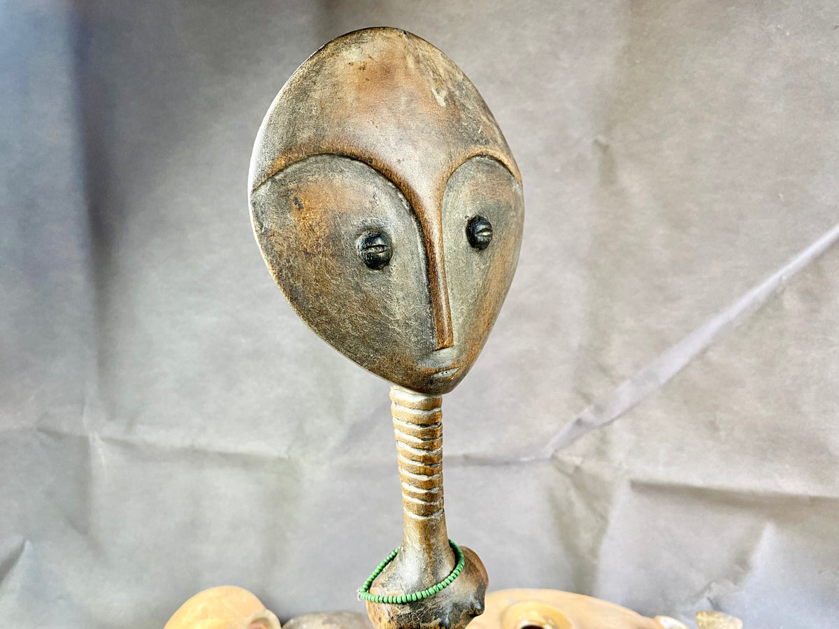 Late 20th Century vintage figure is carved from fine-grained wood, with a natural patina worn smooth with handling. She wears carved neck rings as well as beaded decoration, and a stylised pack on her back. This doll has an well carve face, with other features hinted at rather than explicitly carved. Height 43cm, width at head 11.5 cm.