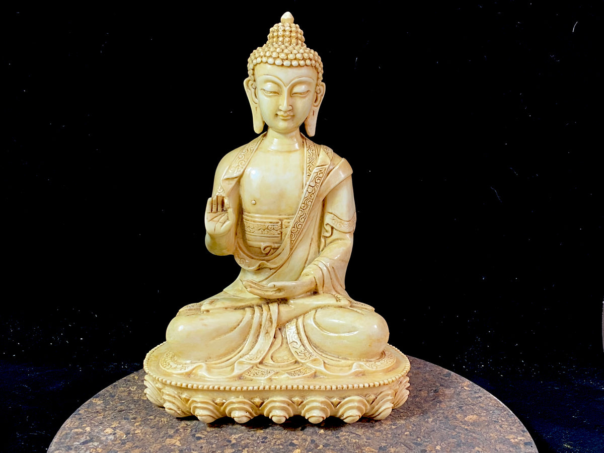Amoghshiddhi Buddha cast in cream coloured resin. His right hand is held up facing outwards in the gesture of protection of fearlessness while his left hand sits in his lap in the gesture of meditation. Cast in solid resin, then hand finished to a very high standard. Measurements: height 26 cm, width 18 cm, depth 12 cm