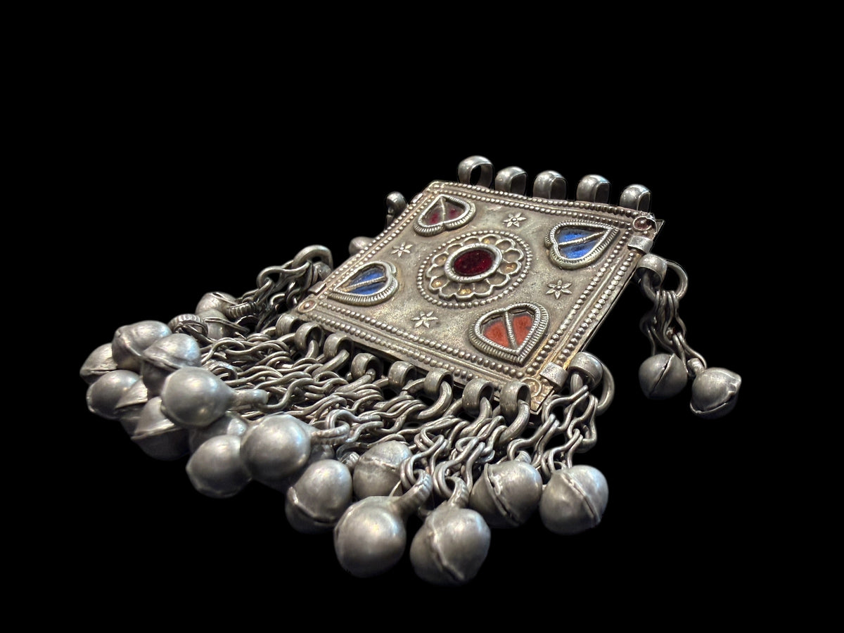 Antique Hunza Valley Uzbek pendant consisting of a flat, double skinned rectangular plaque with a central flower design surrounded by 4 stylised leaves, set with blue and red foil backed glass. Small bells hang in a fringe from the base of the pendant and the sides. Silver and faint gilding. Height 7.4 cm