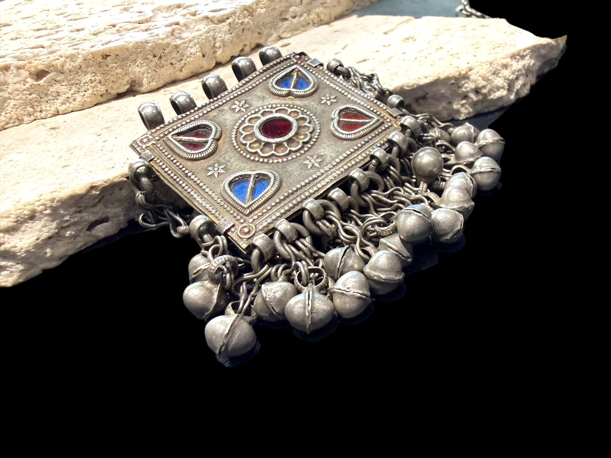 Antique Hunza Valley Uzbek pendant consisting of a flat, double skinned rectangular plaque with a central flower design surrounded by 4 stylised leaves, set with blue and red foil backed glass. Small bells hang in a fringe from the base of the pendant and the sides. Silver and faint gilding. Height 7.4 cm