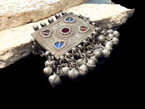 Antique Hunza Valley Uzbek pendant consisting of a flat, double skinned rectangular plaque with a central flower design surrounded by 4 stylised leaves, set with blue and red foil backed glass. Small bells hang in a fringe from the base of the pendant and the sides. Silver and faint gilding. Height 7.4 cm