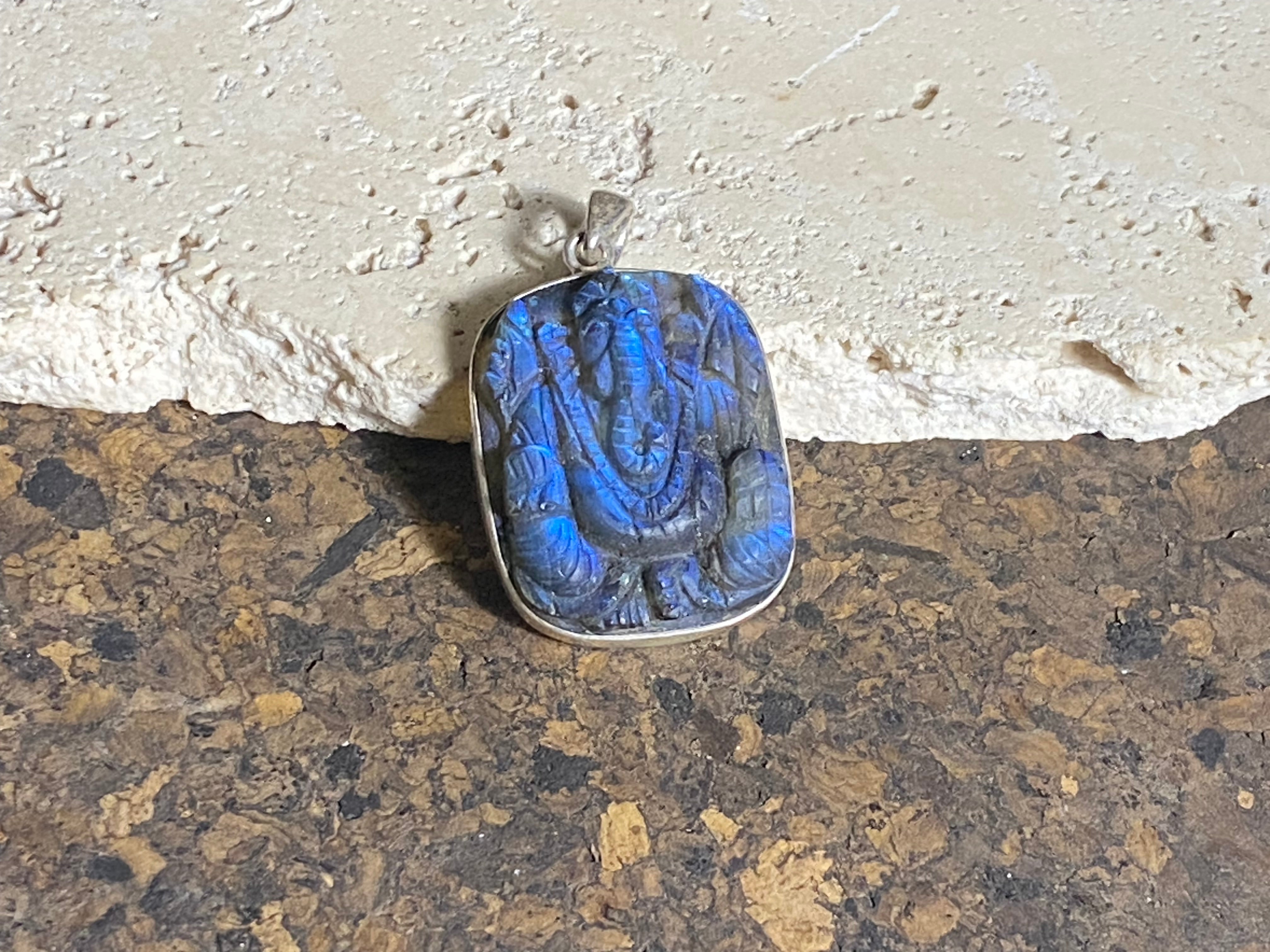 Hand carved square labradorite pendant depicting Ganesh, elephant headed god of wisdom, prosperity and new beginnings. This is a unique and individual piece, hand carved by an artisan in Jaipur, with a light blue luminosity that changes depending on the angle of lighting. Sterling silver frame and bail. The bail is generous enough to allow a large chain or cord to pass through. This unisex labradorite pendant will suit both men and women. Measurements: height 4.7 cm including bail, width 3 cm