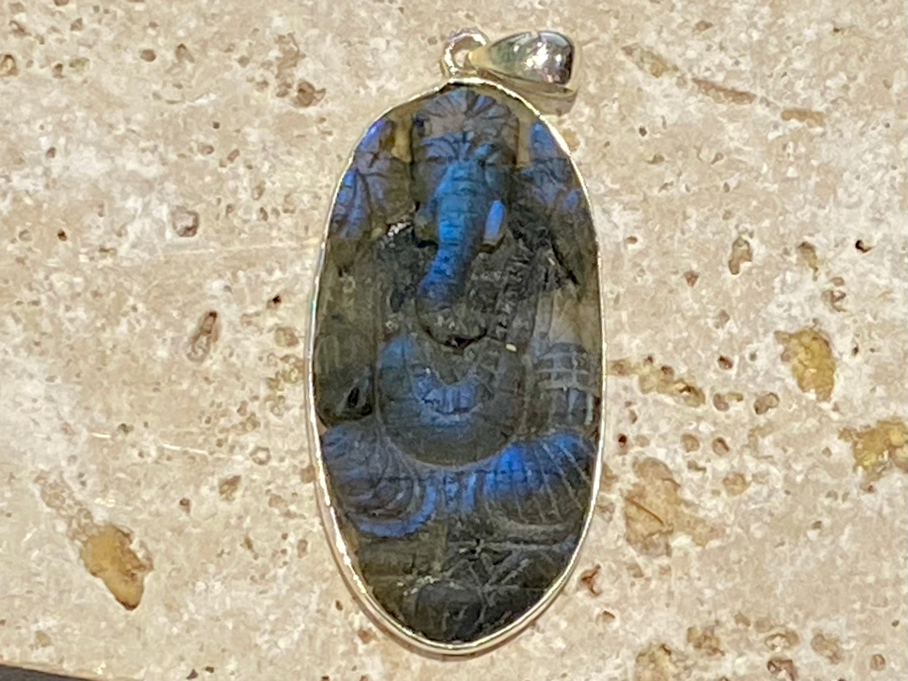 nd carved labradorite pendant depicting Ganesh, elephant headed god of wisdom, prosperity and new beginnings. This is a unique and individual piece, one of our largest, a long oval, with a dark luminosity that comes alive depending on the angle of lighting. Sterling silver frame and bail. The bail is generous enough to allow a large chain or cord to pass through. This unisex labradorite pendant will suit both men and women. Measurements: height 6.2 cm including bail, width 2.7 cm