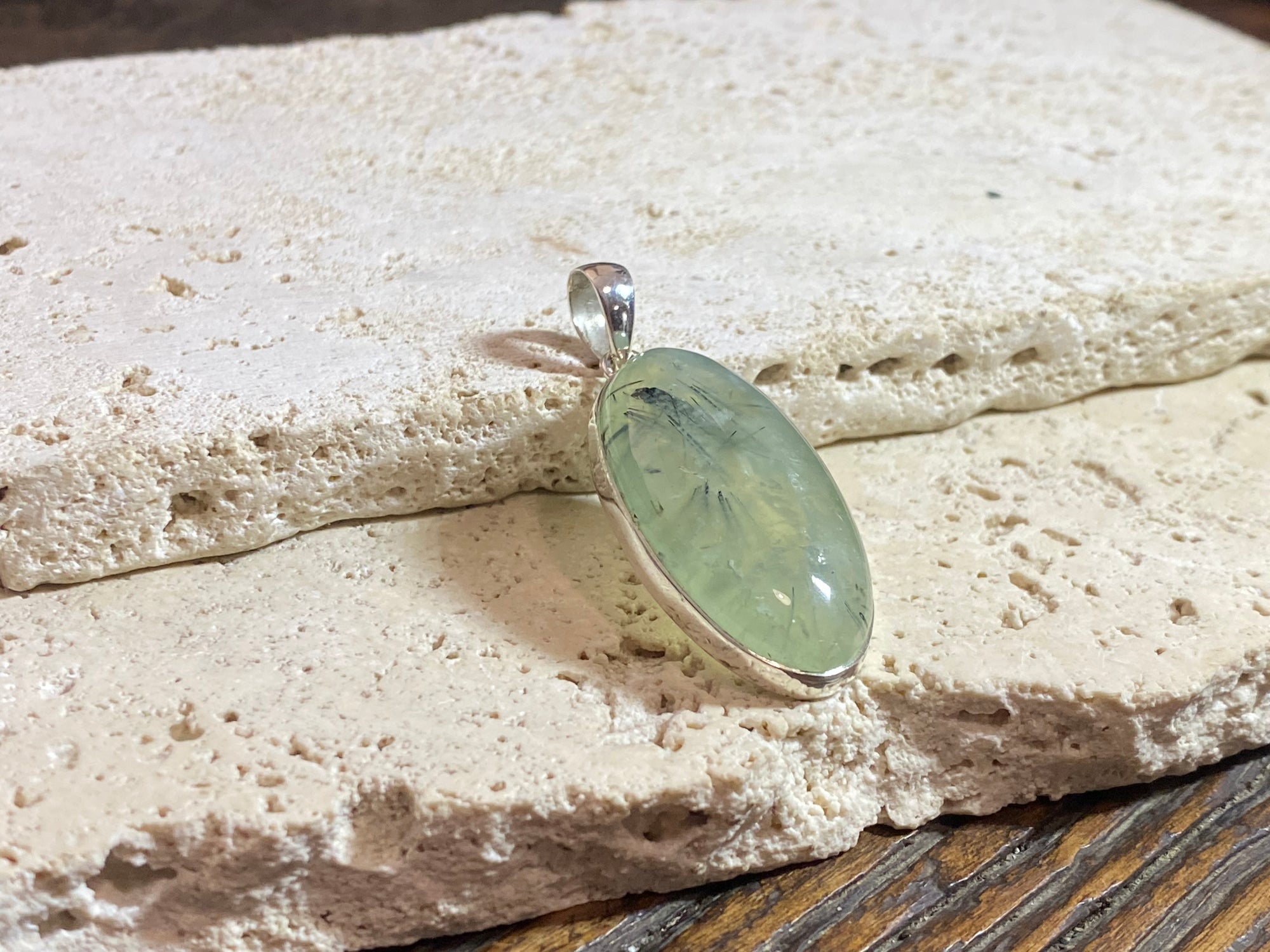 Large oval green prehnite with tourmaline inclusions set in a silver surround with a silver bail that is generous enough to take large chains or cords.