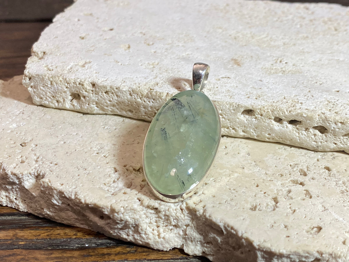Large oval green prehnite with tourmaline inclusions set in a silver surround with a silver bail that is generous enough to take large chains or cords.